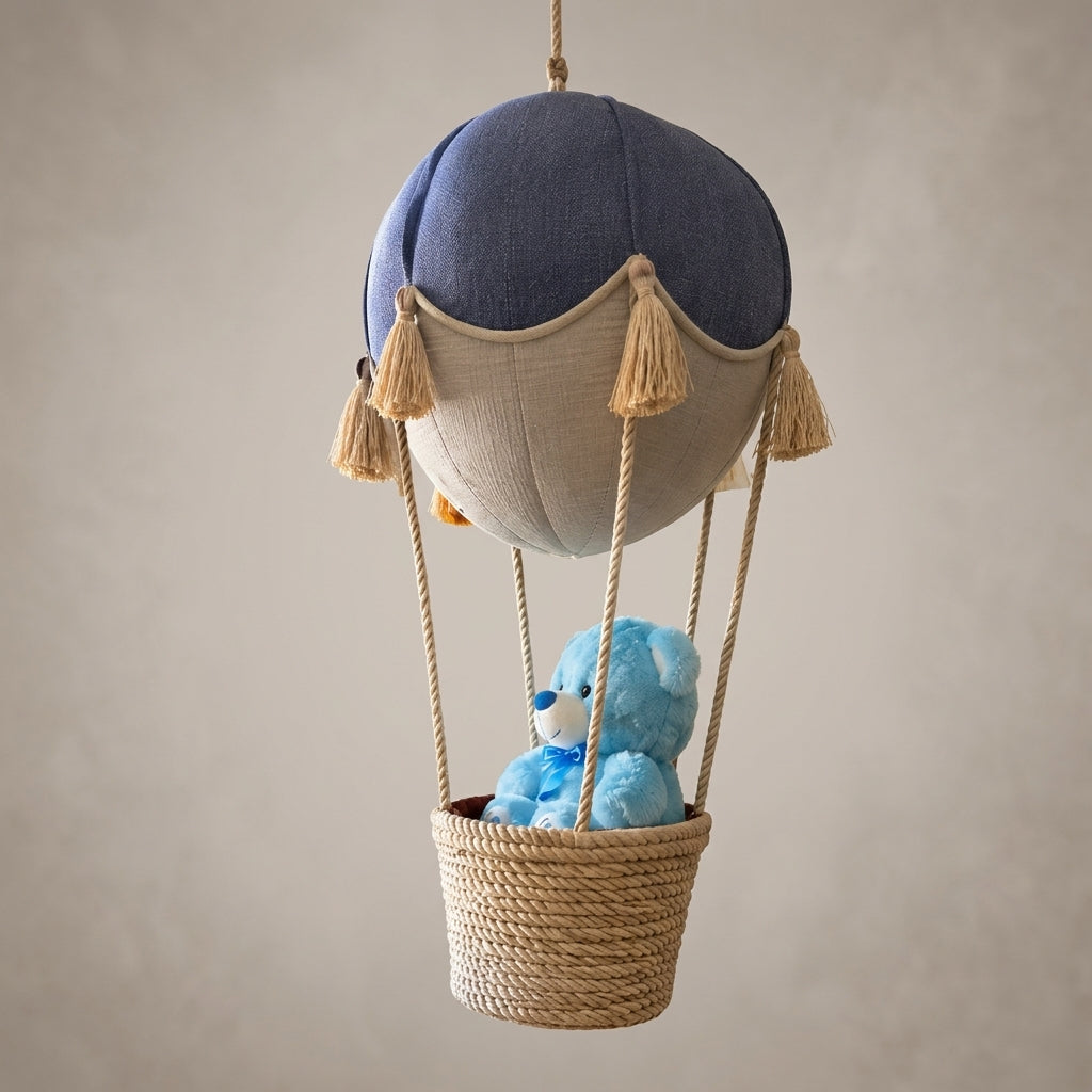 A fabric hot air balloon hanging decoration with a blue and beige balloon body, rope suspension, and a woven basket holding a small blue teddy bear, shown against a plain warm gray background.