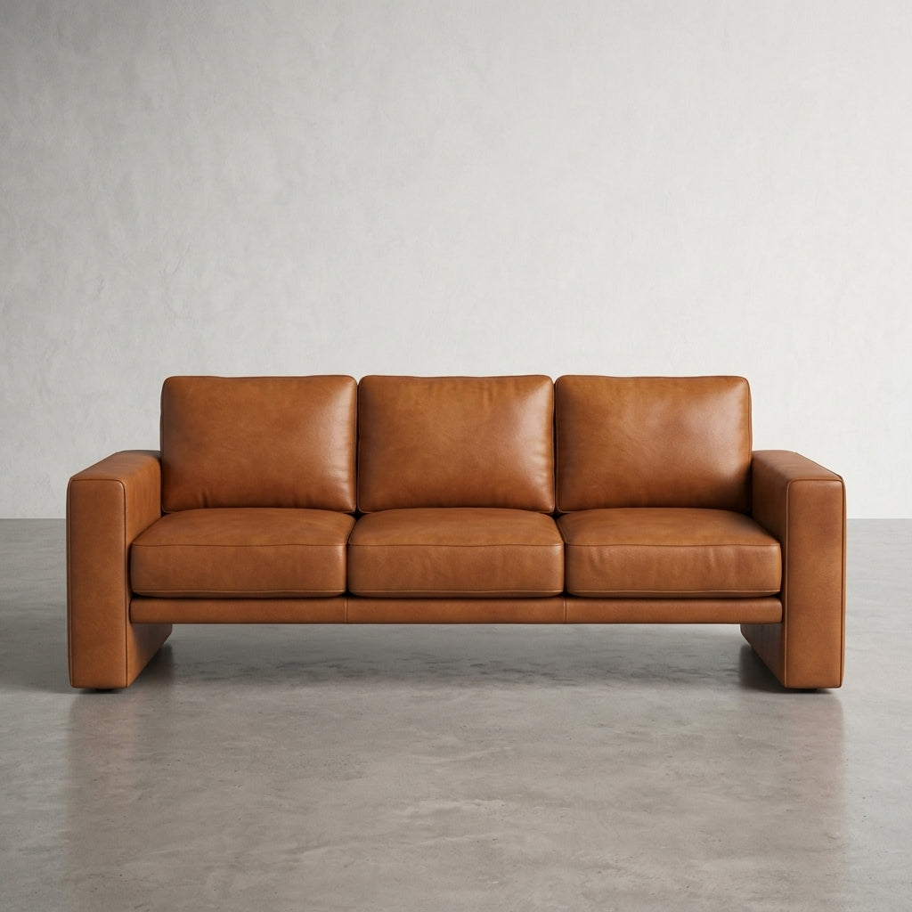 A modern three-seat sofa upholstered in warm tan leather, positioned against a minimalist gray concrete wall and floor.