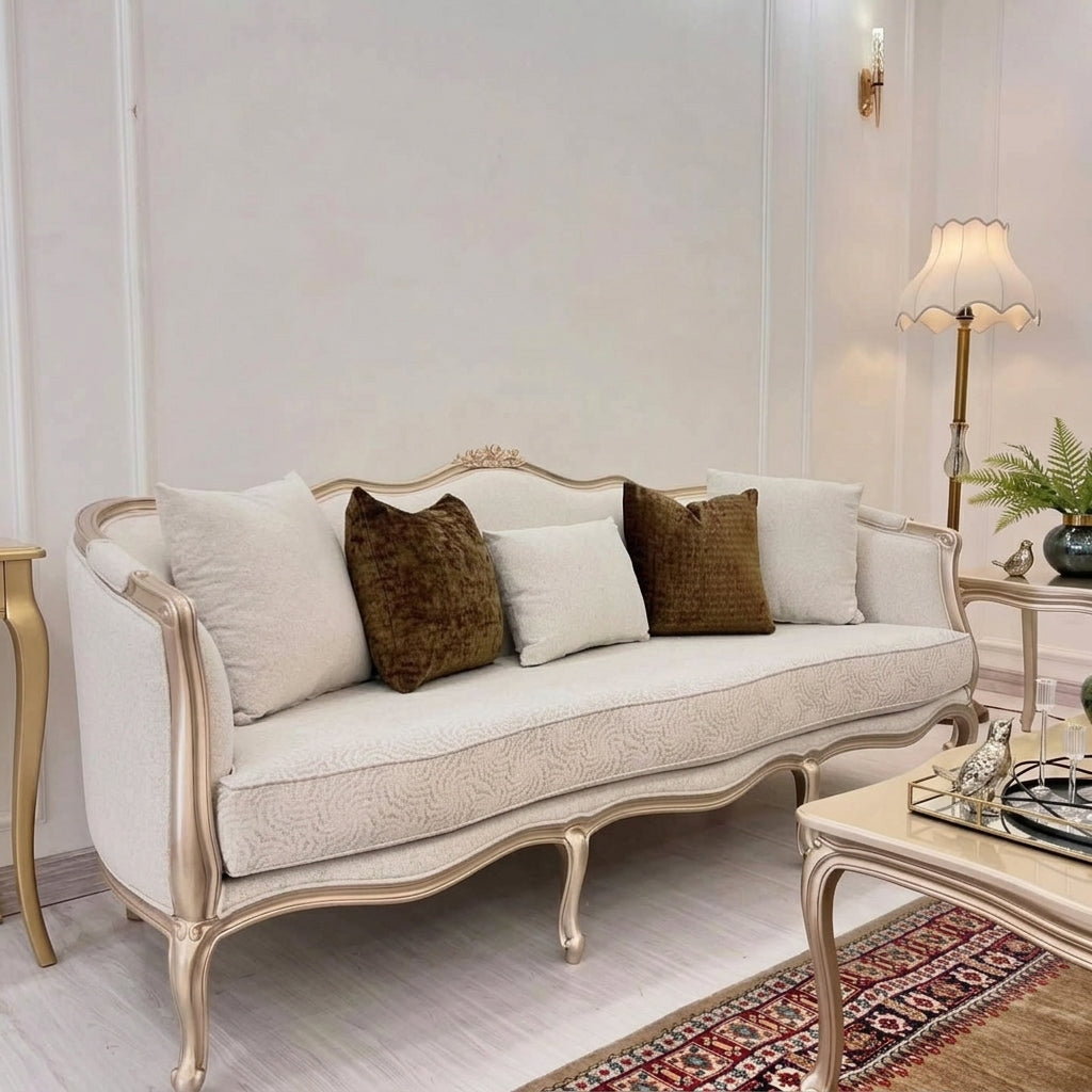 An elegant cream-colored upholstered sofa with ornate gold-toned cabriole legs, styled with four throw pillows (three cream, one textured brown) in a classic living room setting.