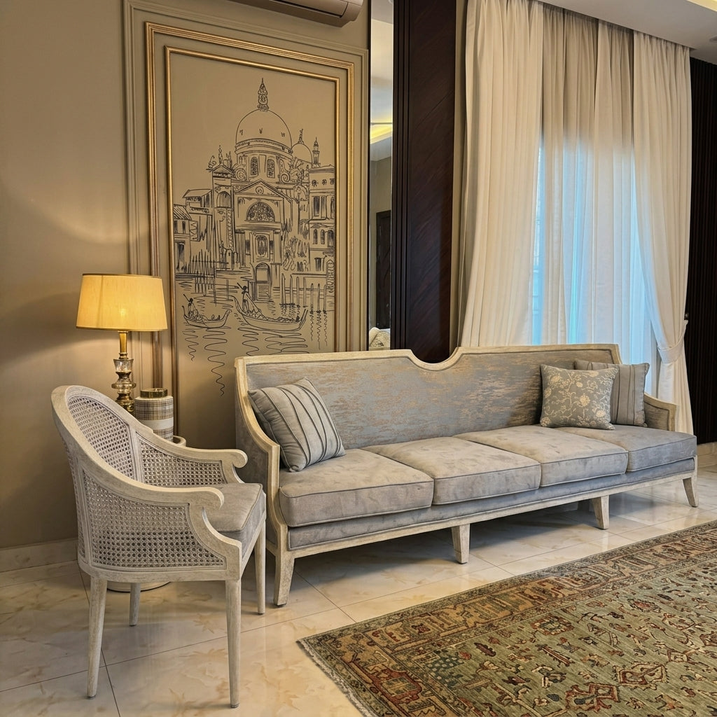 A light blue-gray upholstered sofa with a curved backrest and cream-colored frame, styled in a luxurious living room next to a matching woven armchair, a lit table lamp, and a large framed architectural print.