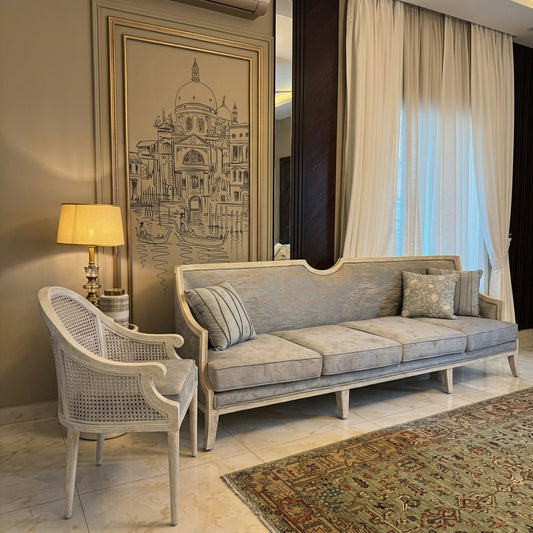 A light blue-gray upholstered sofa with a curved backrest and cream-colored frame, styled in a luxurious living room next to a matching woven armchair, a lit table lamp, and a large framed architectural print.