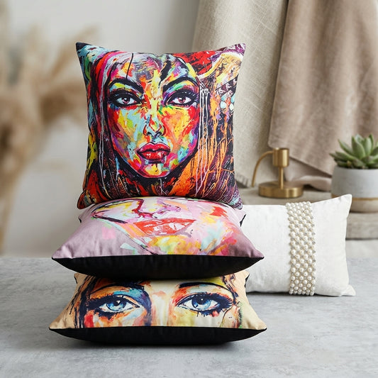 Three decorative throw pillows stacked on a gray surface in a styled interior setting. The top pillow features a vivid multicolor painted face design, the middle pillow shows a pink-toned face illustration, and the bottom pillow displays a close-up of a face with dramatic eyes.