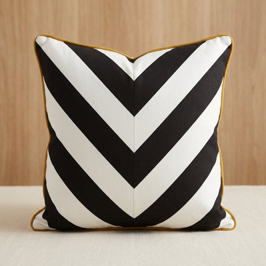A square throw pillow with a bold black and white chevron pattern and a thin gold border, set against a light wood background.