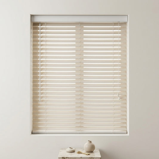 Beige wood blinds with a white frame installed on a window, shown in a clean, minimalist room setting with a small decorative table below.