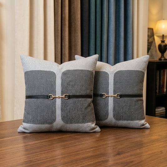 Two square throw pillows in a gray and beige textured fabric with a black leather strap and gold horsebit hardware, displayed on a wooden table in a cozy room setting.