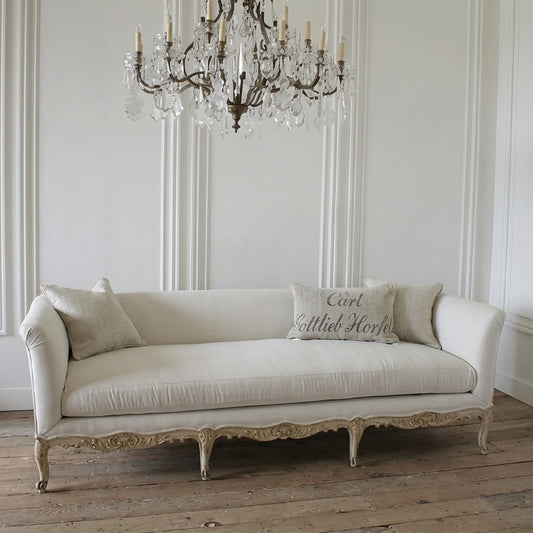 A classic French chaise lounge with a cream linen upholstery and a distressed gold-painted frame, styled in a bright room with a crystal chandelier and wooden floor. One of the throw pillows is embroidered with 'Carl Littled Horry'.