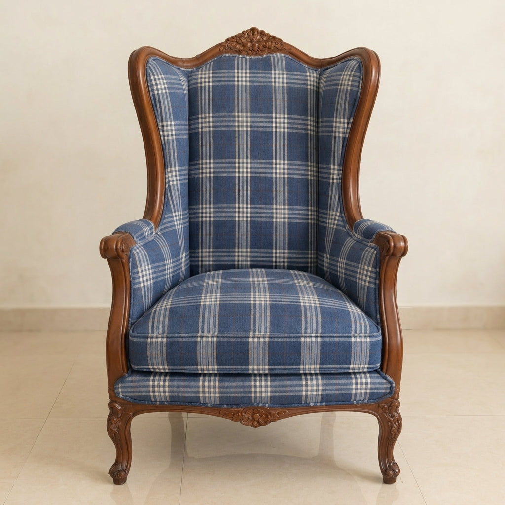 A classic high-back wingback armchair with carved dark brown wooden arms and legs, upholstered in a blue and white plaid fabric, set against a neutral beige wall and tiled floor.
