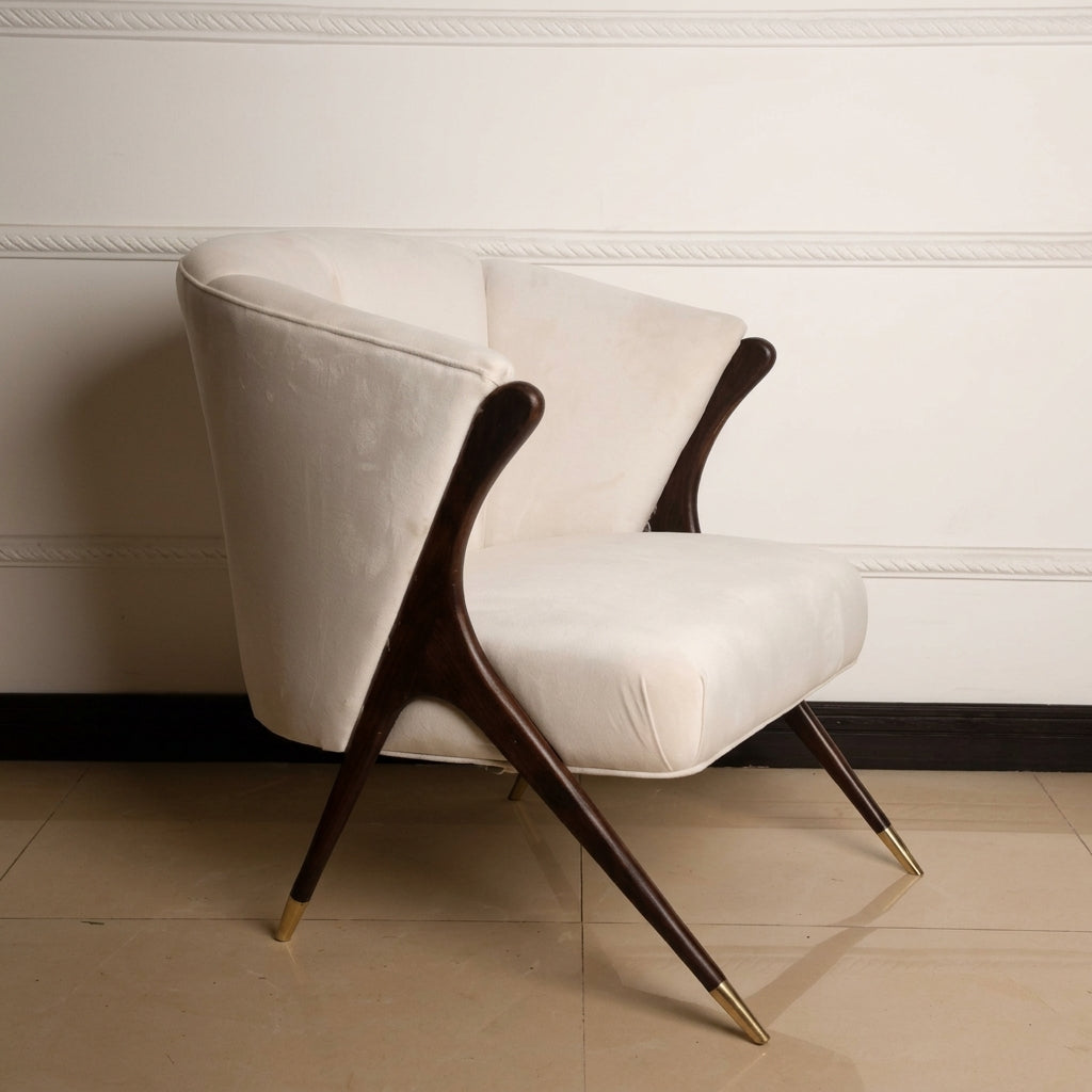 A mid-century modern accent chair with cream velvet upholstery and dark wood legs with gold metal tips, set against a white paneled wall and beige tiled floor.