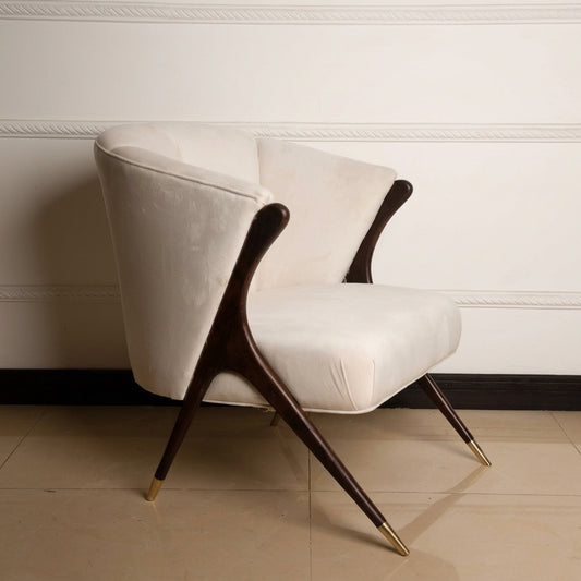 A mid-century modern accent chair with cream velvet upholstery and dark wood legs with gold metal tips, set against a white paneled wall and beige tiled floor.