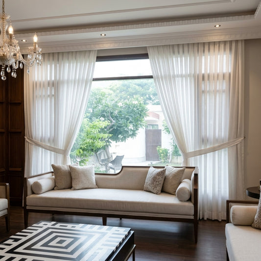 A large window dressed with sheer white curtains and tied back on both sides, showing a light beige upholstered sofa with matching pillows in a styled living room.