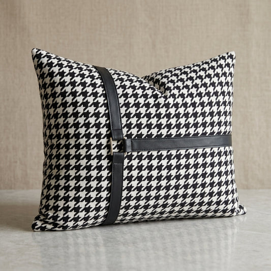 A square throw pillow with a black and white houndstooth pattern, featuring a black faux-leather horizontal strap across the front with a small gold-tone metal accent.