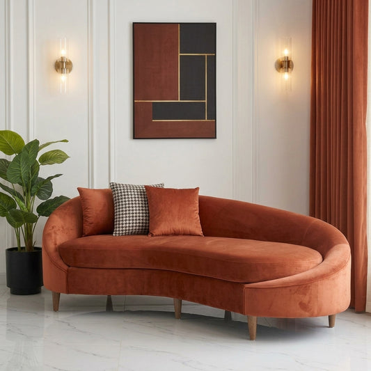 "Modern rust-orange velvet curved chaise lounge sofa with three decorative pillows (two matching velvet, one houndstooth) in a luxury living room setting. The scene includes a marble floor, white wall paneling, a large faux plant, abstract geometric wall art, and brass wall sconces, with terracotta curtains by a large window."
