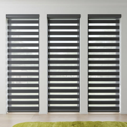 Three dark gray horizontal blind window treatments installed on tall windows in a bright room with light walls and a pale wood floor. A lime green rug is partially visible at the bottom.