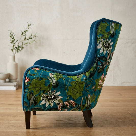 A wingback armchair upholstered in teal velvet with a botanical floral pattern on the outer arms and back, shown in a styled room setting with a light wood floor and textured wall.