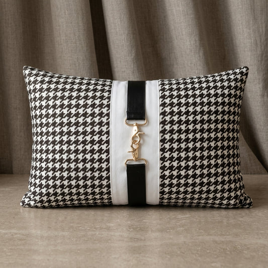 A rectangular lumbar throw pillow with a black and white houndstooth pattern, featuring a central vertical white stripe with black leather-like trim and a gold-tone metal clasp closure, set against a neutral draped fabric backdrop.