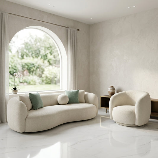 A modern curved cream bouclé sofa with two sage green pillows is shown in a bright, minimalist living room with a matching cream armchair, textured walls, and a large arched window.
