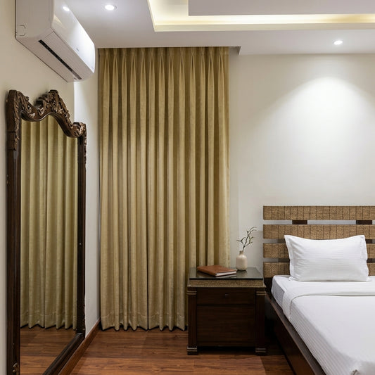 A set of floor-length beige curtains hanging in a bedroom next to a wooden floor mirror and a bed with white linens.